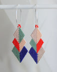 A pair of handmade beaded geometric dangle earrings hanging from silver hoop earrings. The earrings feature a stacked diamond pattern in sage green, light peach, bright red, and royal blue beads, with the peach and green sections textured using a seed bead chevron pattern.
