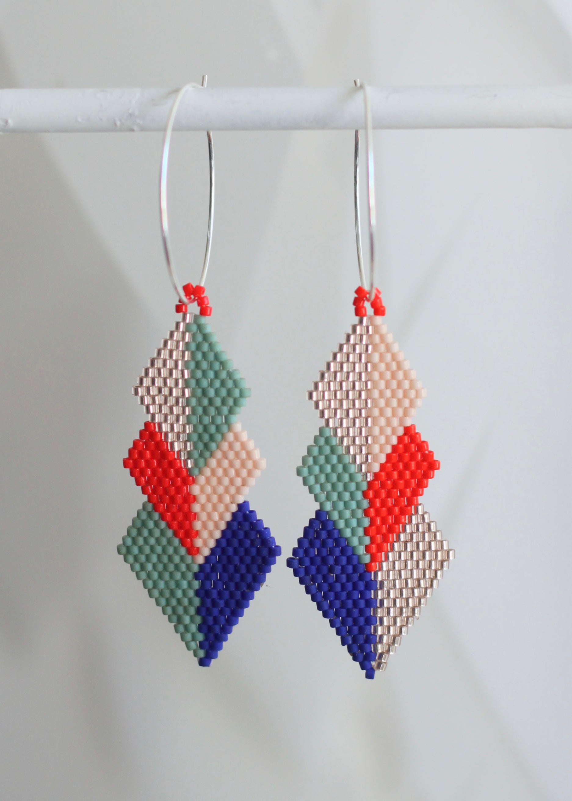 A pair of handmade beaded geometric dangle earrings hanging from silver hoop earrings. The earrings feature a stacked diamond pattern in sage green, light peach, bright red, and royal blue beads, with the peach and green sections textured using a seed bead chevron pattern.